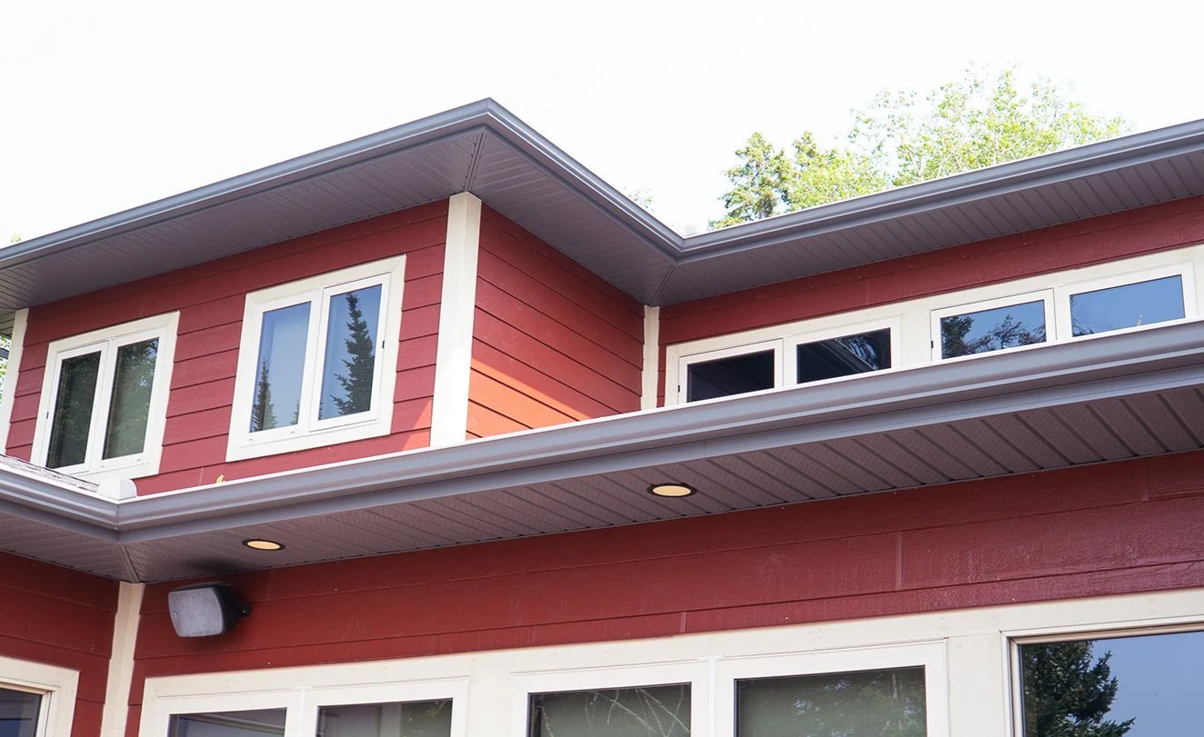 Close-up of a two-story house with red siding, white trim, multiple large windows, and a gray roof, surrounded by trees.