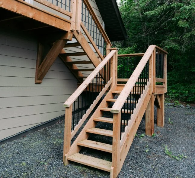 Wooden outdoor staircase with black metal railings leading to a deck attached to a house, surrounded by trees and gravel ground.