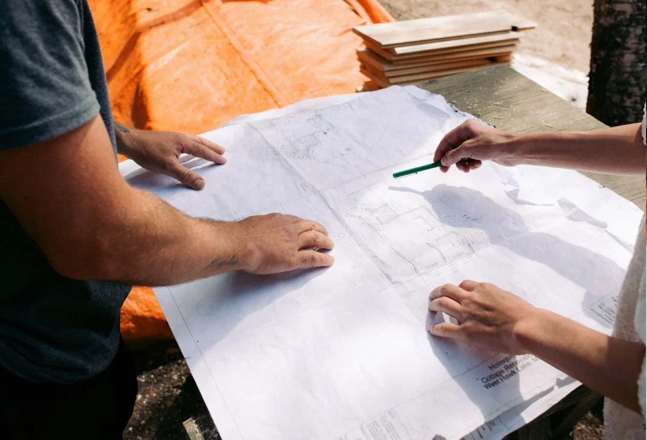 Two people reviewing architectural blueprints outdoors, with one person pointing at a section of the blueprints, on a table with a stack of paper nearby.