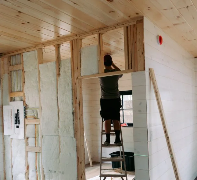A person wearing a gray shirt, black shorts, and a black cap standing on a ladder, working on the upper part of a wooden wall in an interior construction site, with insulation and electrical panel visible.