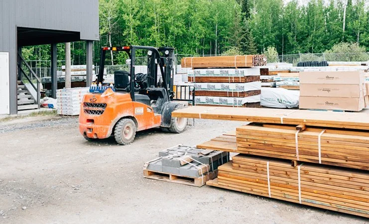 Construction site with a forklift and stacks of lumber and building materials outside a building.