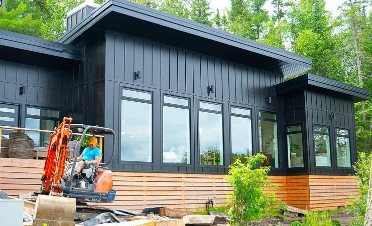 Modern black and wood house with large windows, construction worker operating an excavator outside, surrounded by green trees.