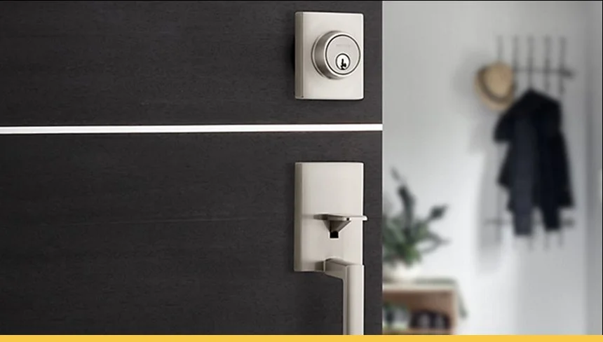 Close-up of a modern black door with a white doorknob and lock, with a blurred background showing a wall with hooks, hanging coats, and a plant.