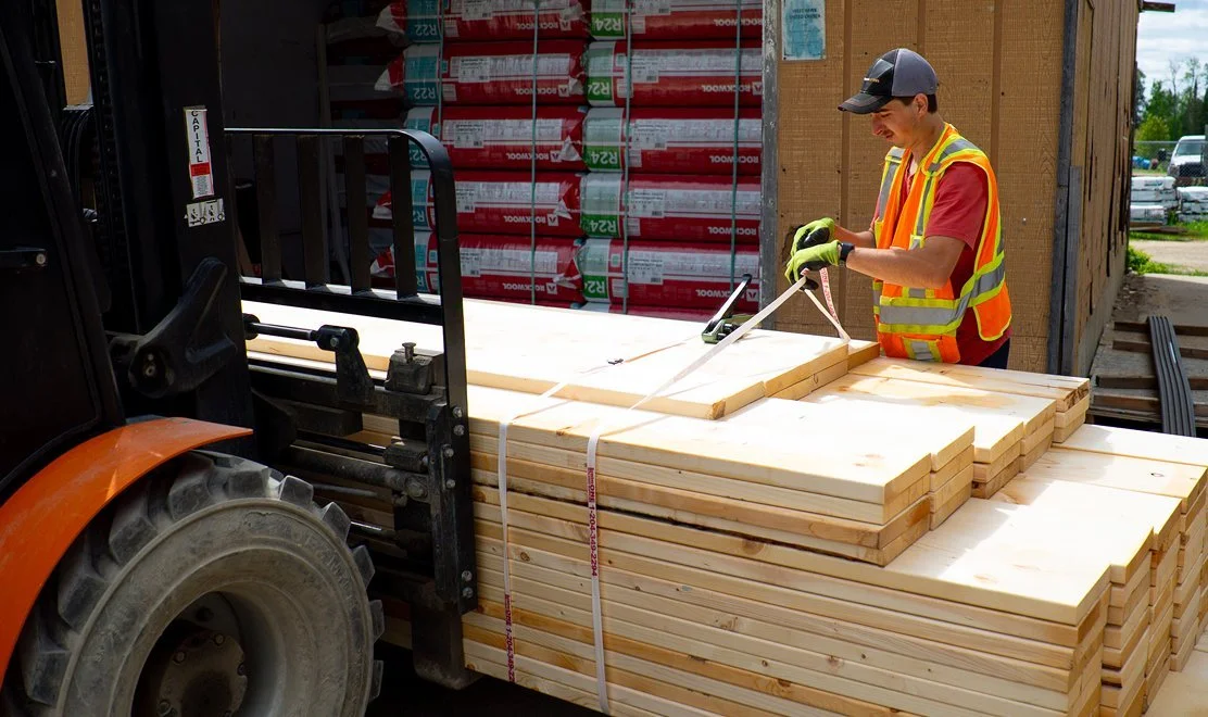A man in a safety vest and cap securing lumber with a tape measure next to a forklift loaded with wooden planks, in an outdoor construction materials yard.