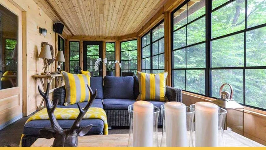 Cozy sunroom with large windows showing green trees outside, wooden walls and ceiling, dark wicker sofa with yellow and gray pillows, candles on the table, antler decor, and a small side table with a basket.