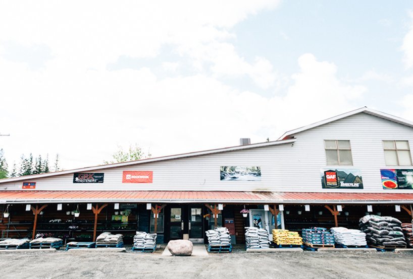 Storefront with signs and stacked items outside under a wooden awning, building has a metal roof, partly cloudy sky