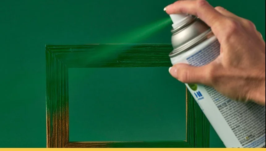 A person spraying green spray paint on a rectangular picture frame against a green background.
