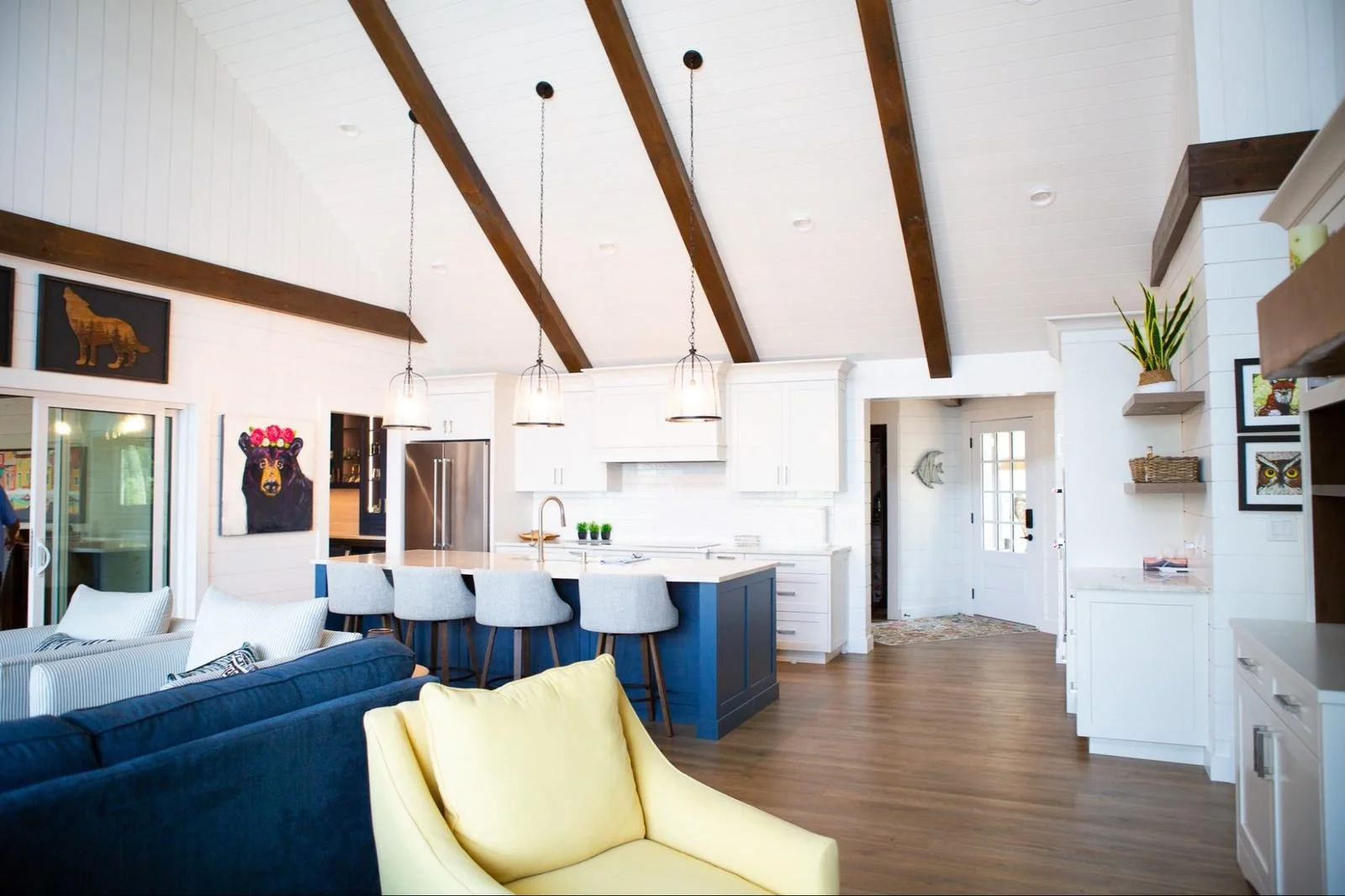 Bright open-concept living room and kitchen with white walls, wooden ceiling beams, gray and yellow armchairs, white kitchen cabinets, gray island with barstools, and decorative art and plants.