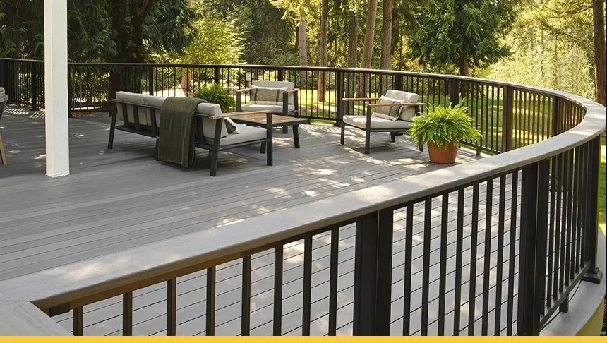 View of a spacious outdoor deck with patio furniture, including sofas and chairs, surrounded by a black railing, with trees and greenery in the background.