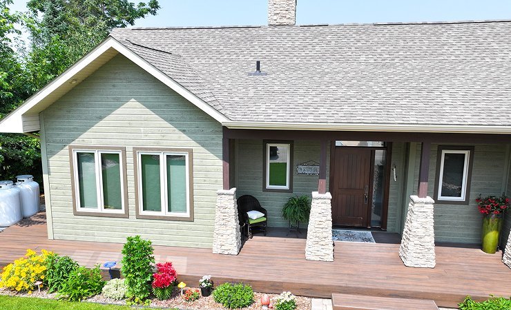Front view of a house with a wooden porch, three windows, a wooden door, and decorative stone columns. There are potted plants and flowers on the porch, and a lawn with colorful flowers in front.