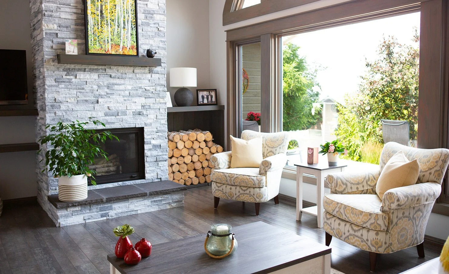 Living room with a stone fireplace, logs, armchairs, large windows, and plants.