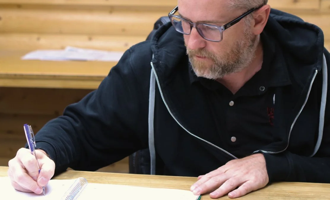 A man with glasses, a beard, and a black hoodie writing in a notebook at a wooden desk.