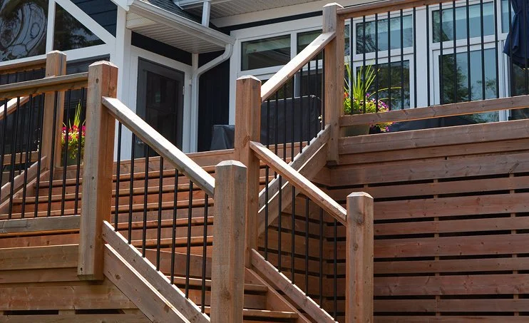 Wooden outdoor stairs with black metal railings leading to a patio with flower pots and a glass door, attached to a modern building.