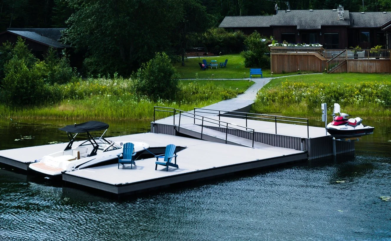 A lakeside dock with two boats, one covered, and two blue Adirondack chairs, leading to a grassy yard with trees and a house in the background.
