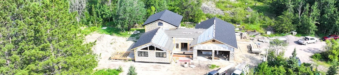 A house under construction surrounded by trees, with construction materials, multiple cars, and a driveway visible.