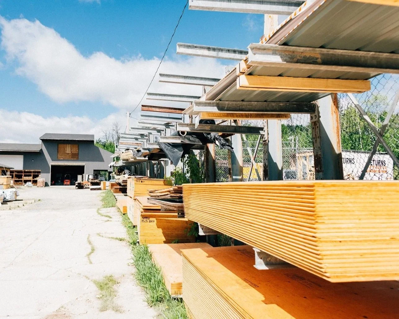 Outdoor lumber yard with stacks of wooden planks on metal racks under a partly cloudy sky.