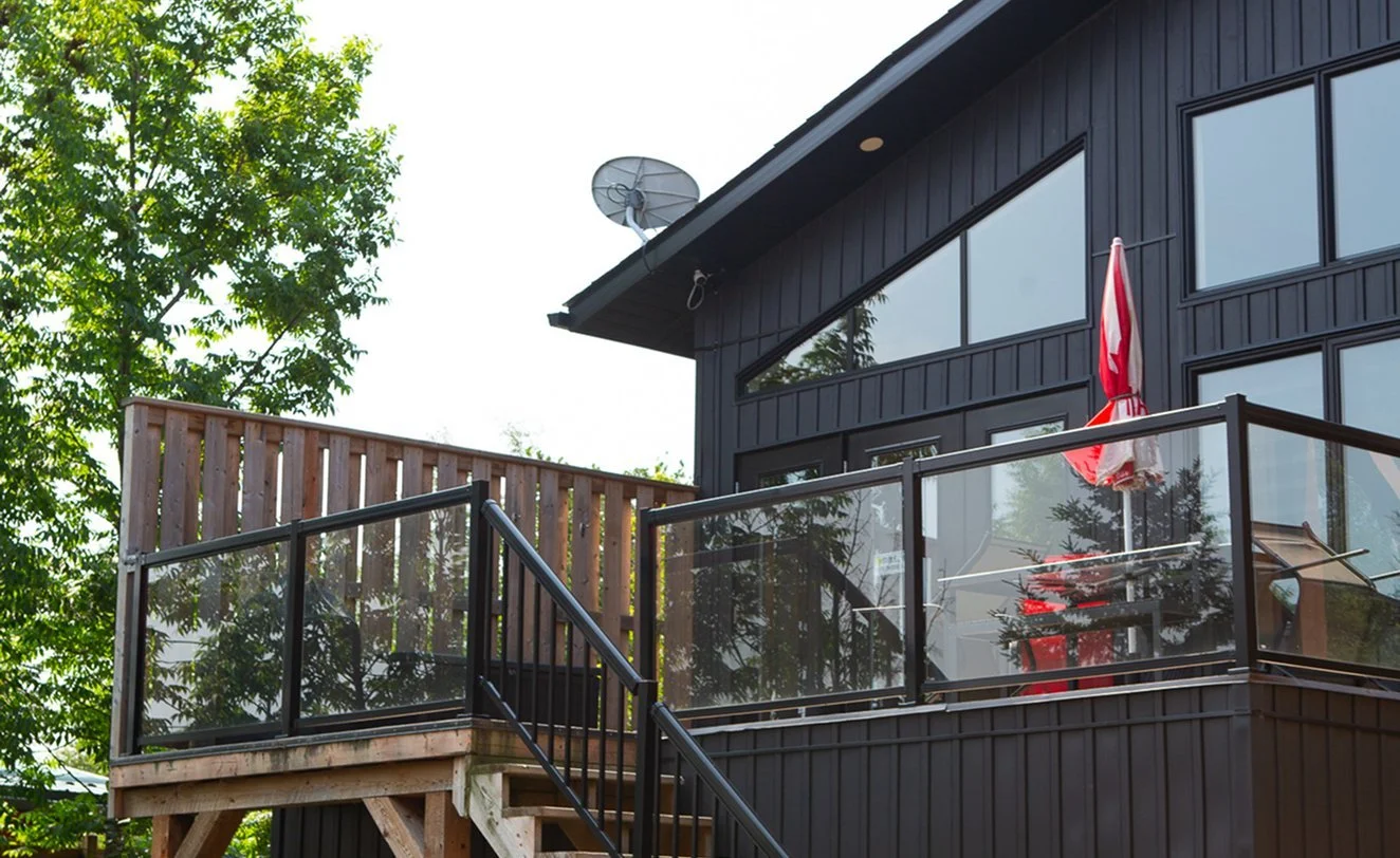 A black modern house with large glass windows and a balcony with a glass railing. An American flag and a red patio umbrella on the balcony. A satellite dish attached to the roof. Green trees behind the house.