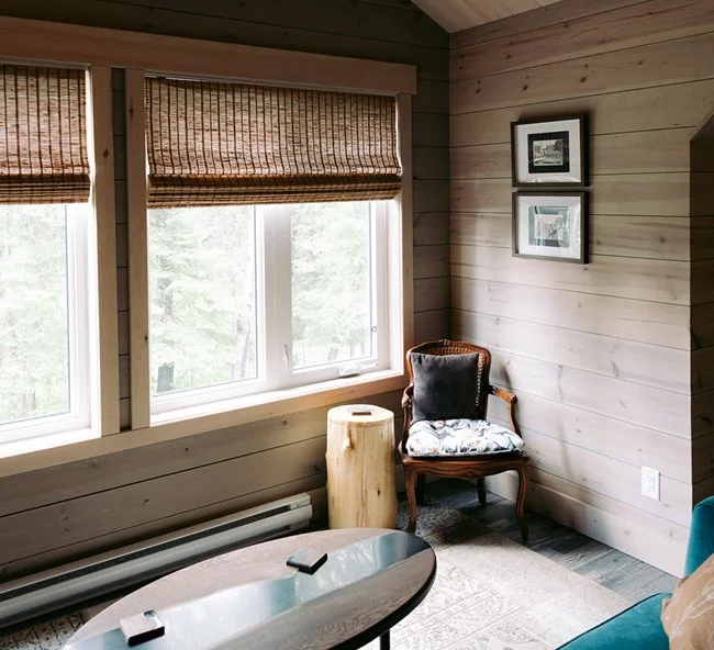 Corner of a cozy room with a window, wooden wall paneling, a vintage armchair with a cushion, a side table, and framed pictures on the wall.