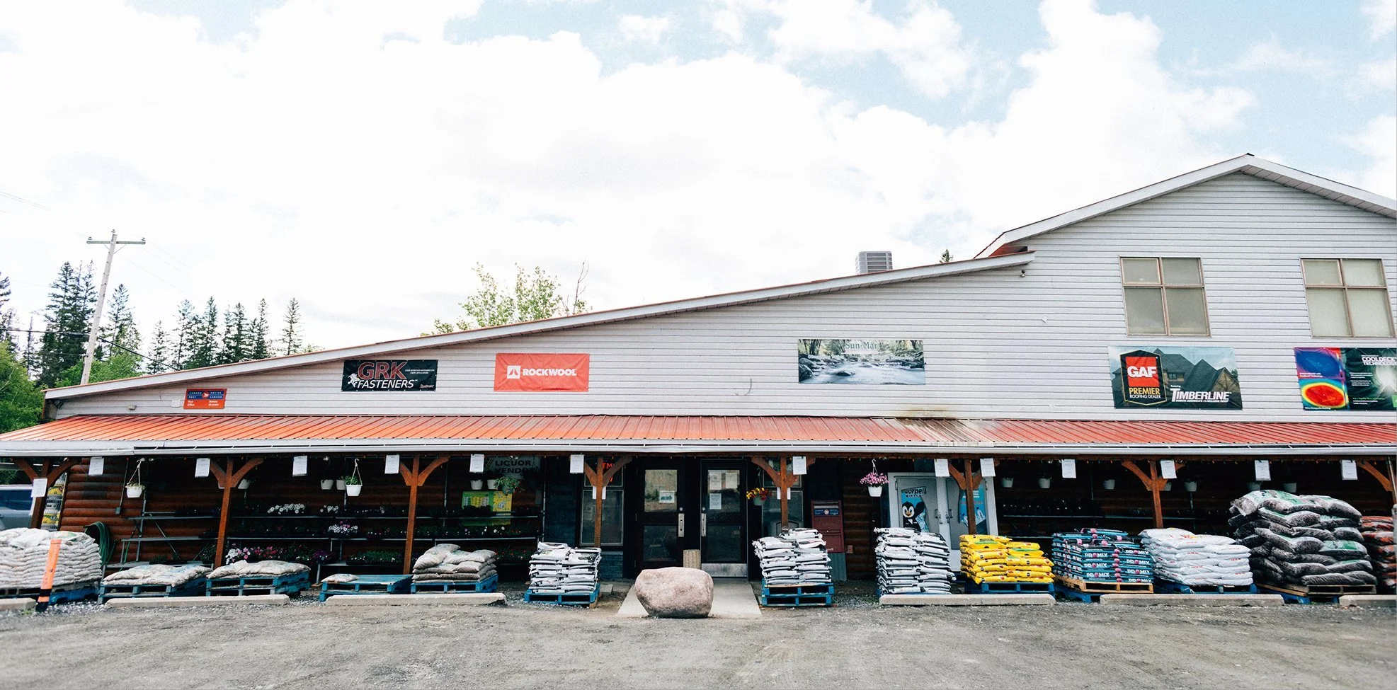 View of the front of a hardware store with a wood and metal exterior, displaying various advertisements for brands like GAF, Timberline, and Rockwool. Outside are bags of soil, fertilizer, and gardening supplies, with a gravel parking area in front.