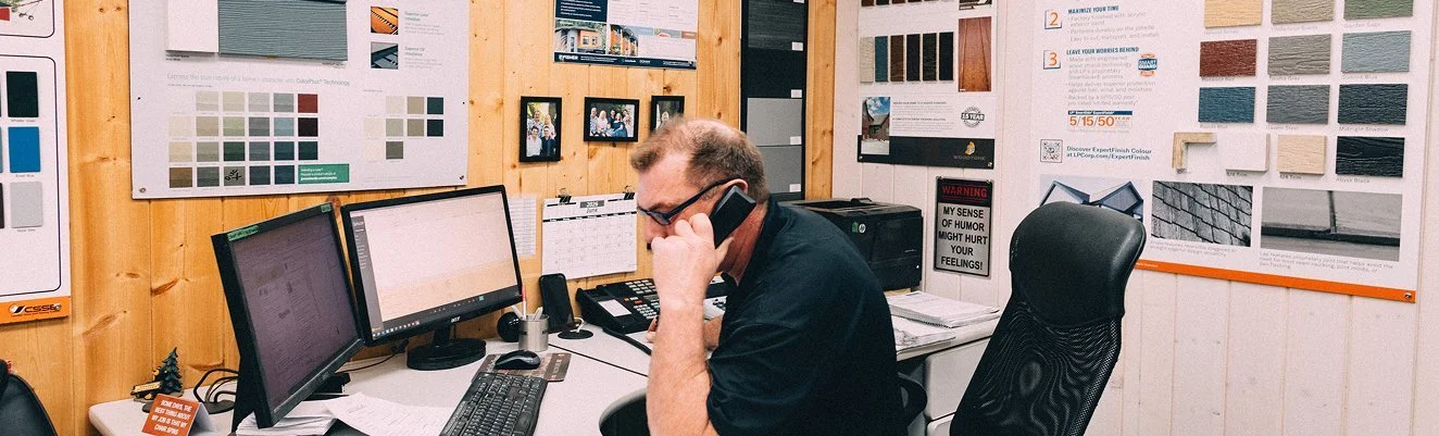 Man in glasses talking on the phone in an office with dual computer monitors, a telephone, and various papers on the desk, with wood-paneled walls covered in posters and framed pictures.