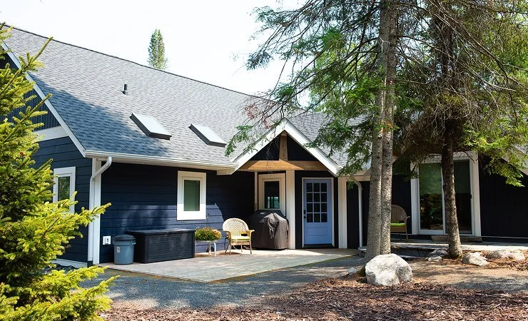 A house with a blue exterior, a shingled roof, and a porch area. There are patio chairs, a grill, and trees surrounding the house.