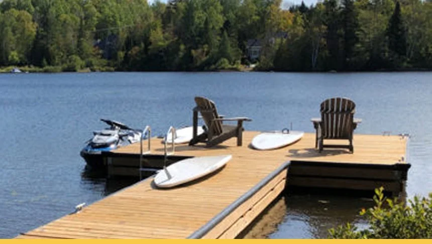A wooden dock extending into a lake, with two Adirondack chairs, two paddleboards, and a kayak, set against a backdrop of trees and clear sky.