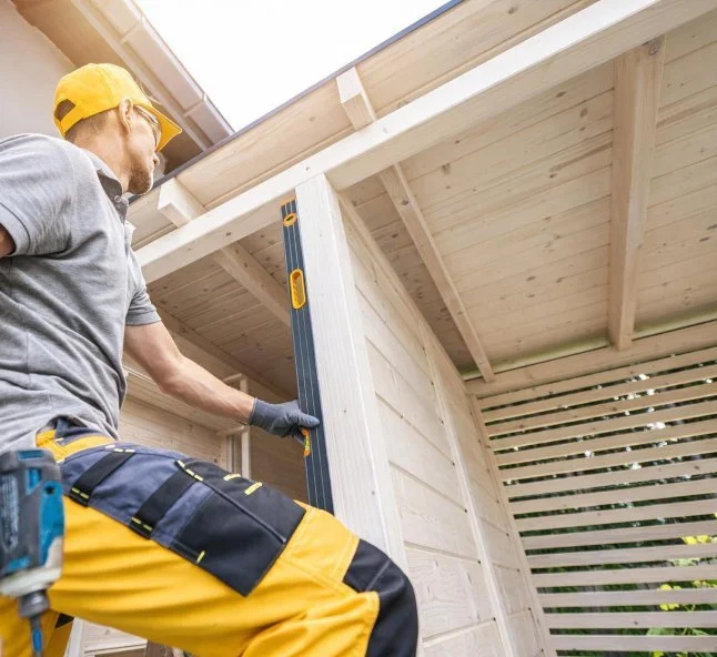 A man wearing a yellow cap, gray shirt, and yellow work pants measuring a wooden corner of a new outdoor shed under construction.