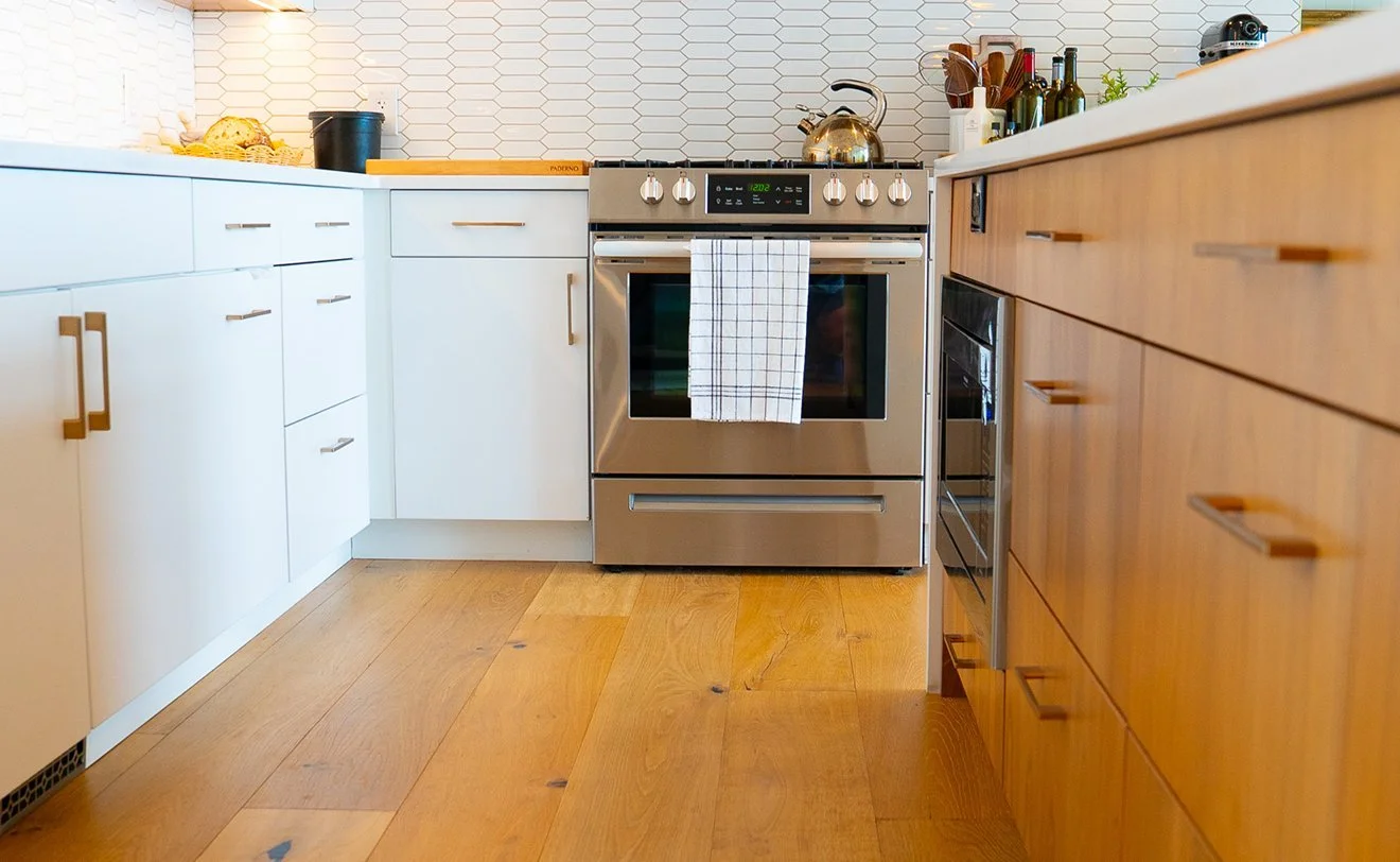 Kitchen with white cabinets on the left, a stainless steel stove in the center with a towel hanging on the oven handle, and wooden cabinets on the right, with various bottles and kitchen utensils on the countertop.
