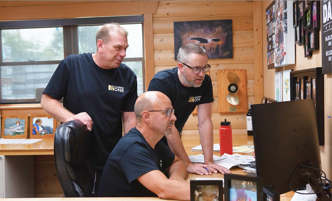 Three men working together in a wood-paneled office space, looking at a computer screen. One man is seated, and two are standing beside him. The room has framed photos, artwork, and a wall-mounted speaker.