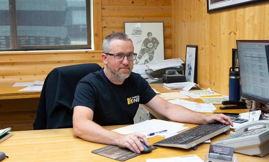 A man with gray hair and beard, wearing glasses and a black T-shirt, sitting at a cluttered office desk with papers, a keyboard, and two monitors in a wood-paneled office with a large window.