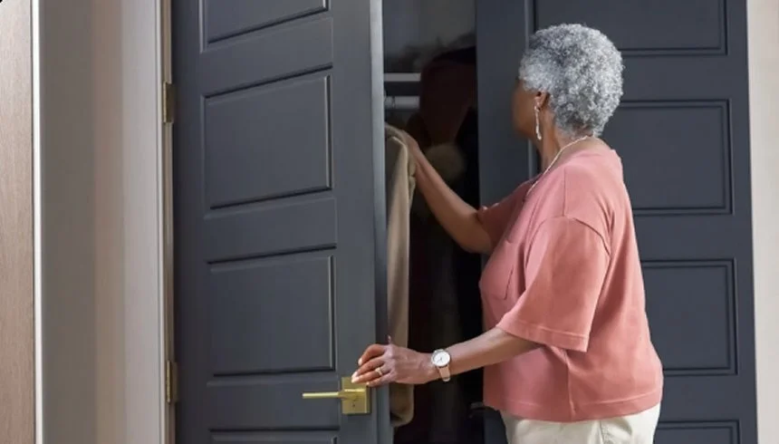 An elderly woman with gray curly hair, wearing a pink blouse and white pants, opening a dark gray closet door to look inside.