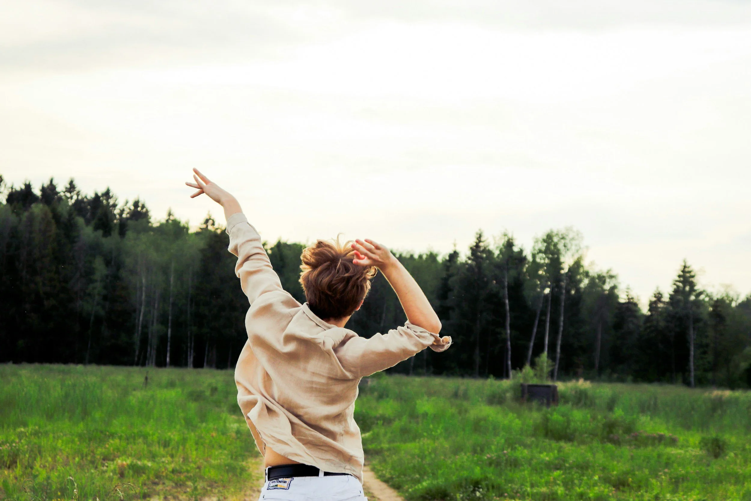 A person with short brown hair, wearing a beige jacket, standing in a grassy field with trees in the background, raising both arms up towards the sky.