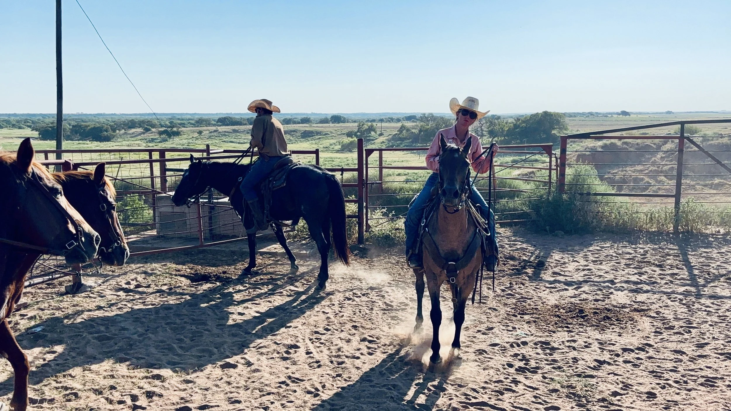 Two women riding horses in an outdoor corral with open fields and blue sky in the background, wearing hats and sunglasses.