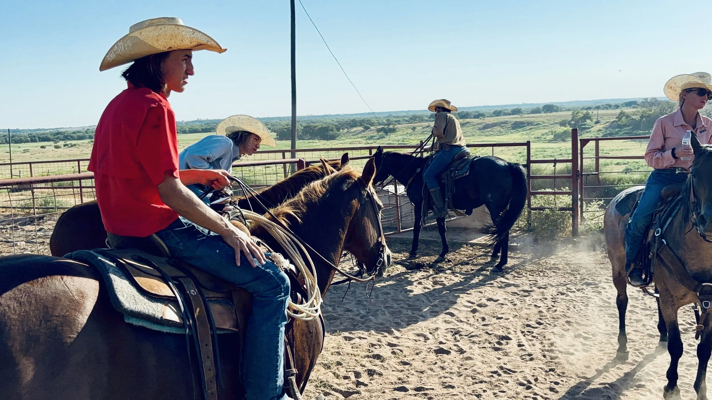 Group of people horseback riding in an open field, wearing cowboy hats, with a rural landscape in the background.