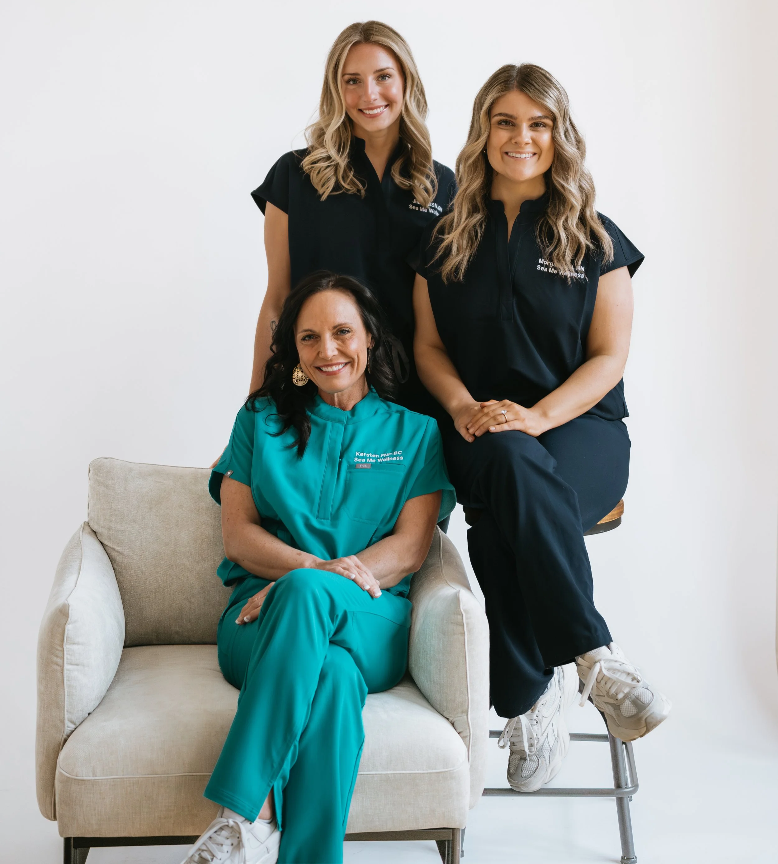 Three Sea Me Wellness team members smiling together, two standing in dark scrubs and one seated in teal scrubs.