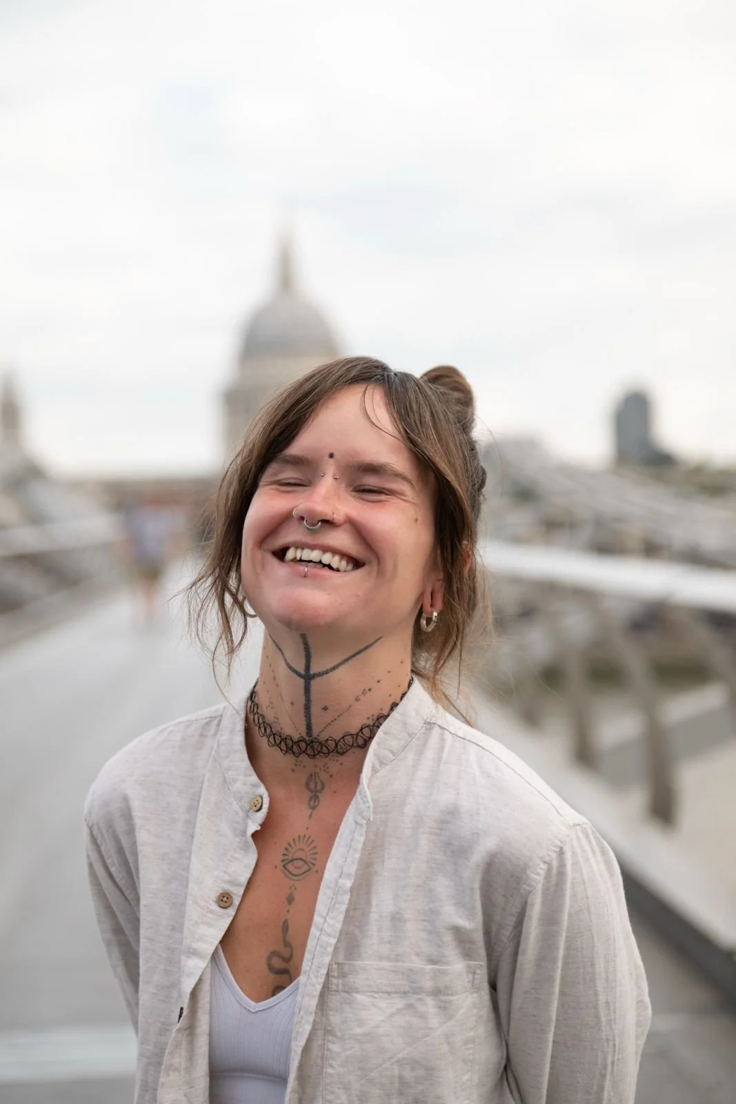 A smiling young woman with tattoos and piercings is standing outdoors in front of a cityscape with a blurred domed building in the background.