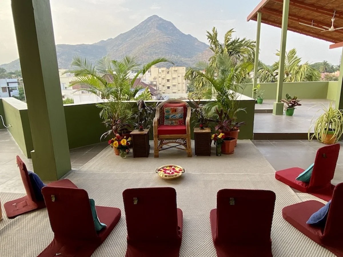 Outdoor balcony with red chairs, potted plants, a mountain view in the background, and a central chair with floral decorations on a carpeted floor.