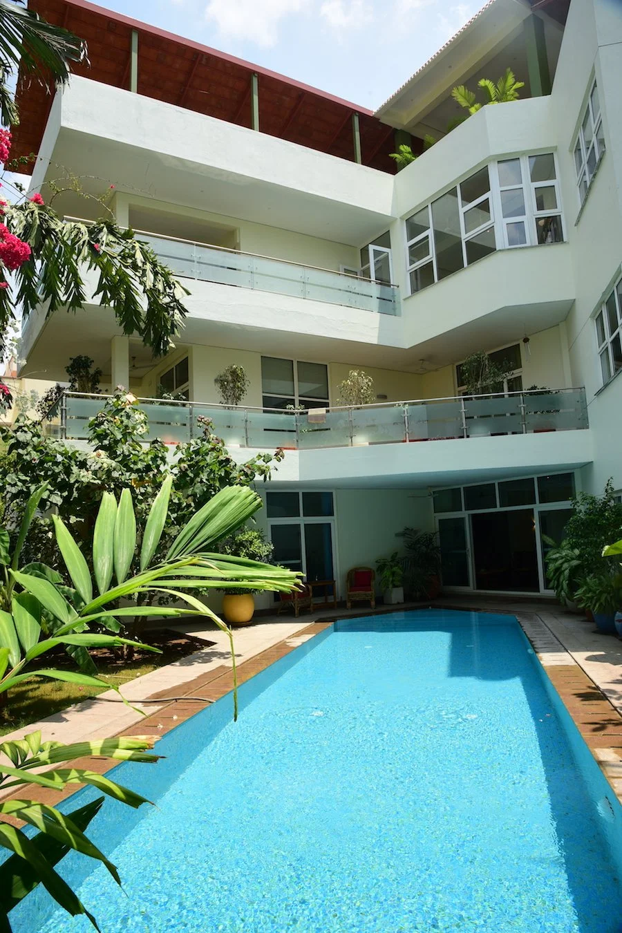 A view of a modern multi-story building with white walls and large windows, surrounding a blue swimming pool in a courtyard filled with green plants and outdoor furniture.