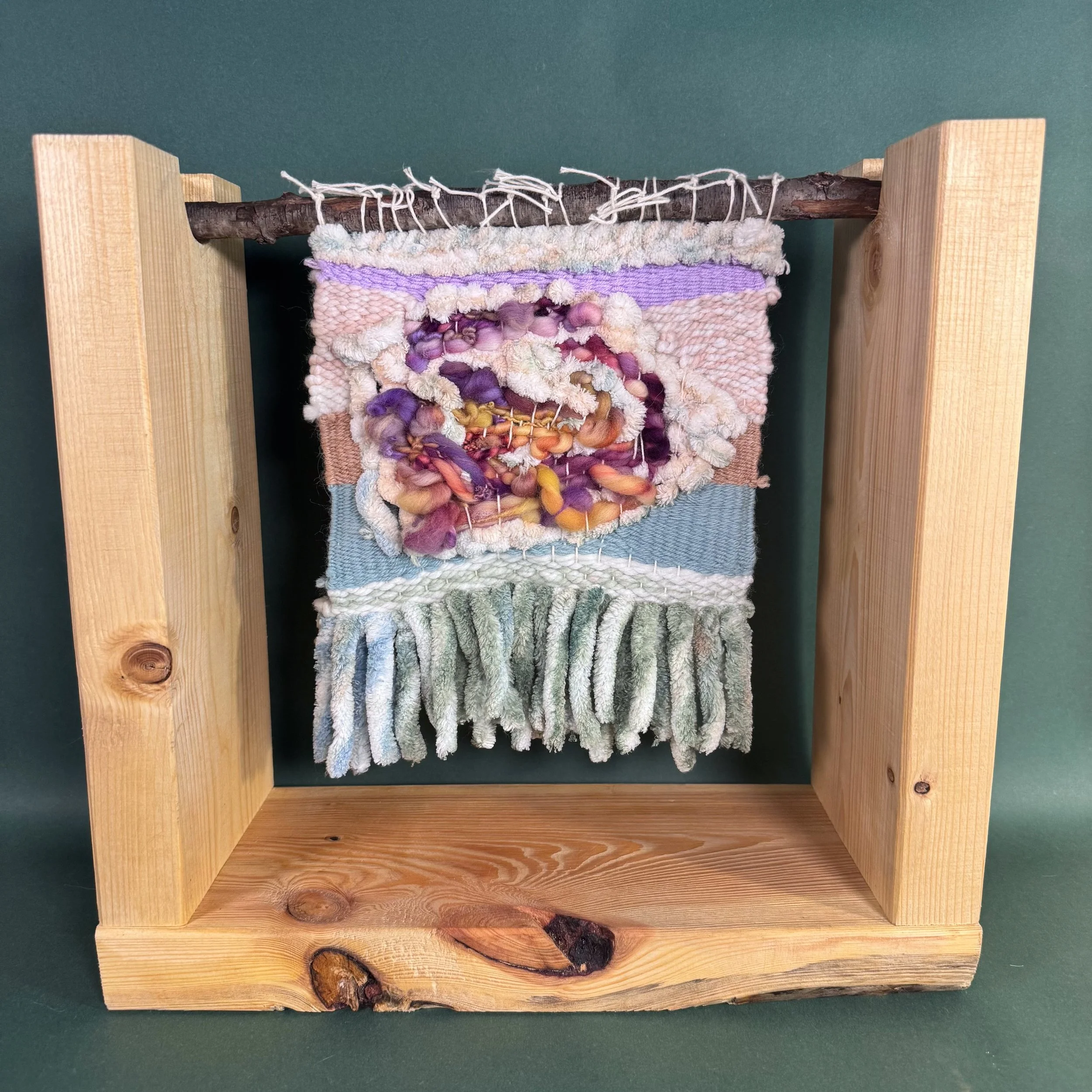 A woven wall hanging with a wooden frame, featuring various textures and colors of yarn and fabric, including shades of purple, pink, orange, and green, supported by a horizontal wooden stick.
