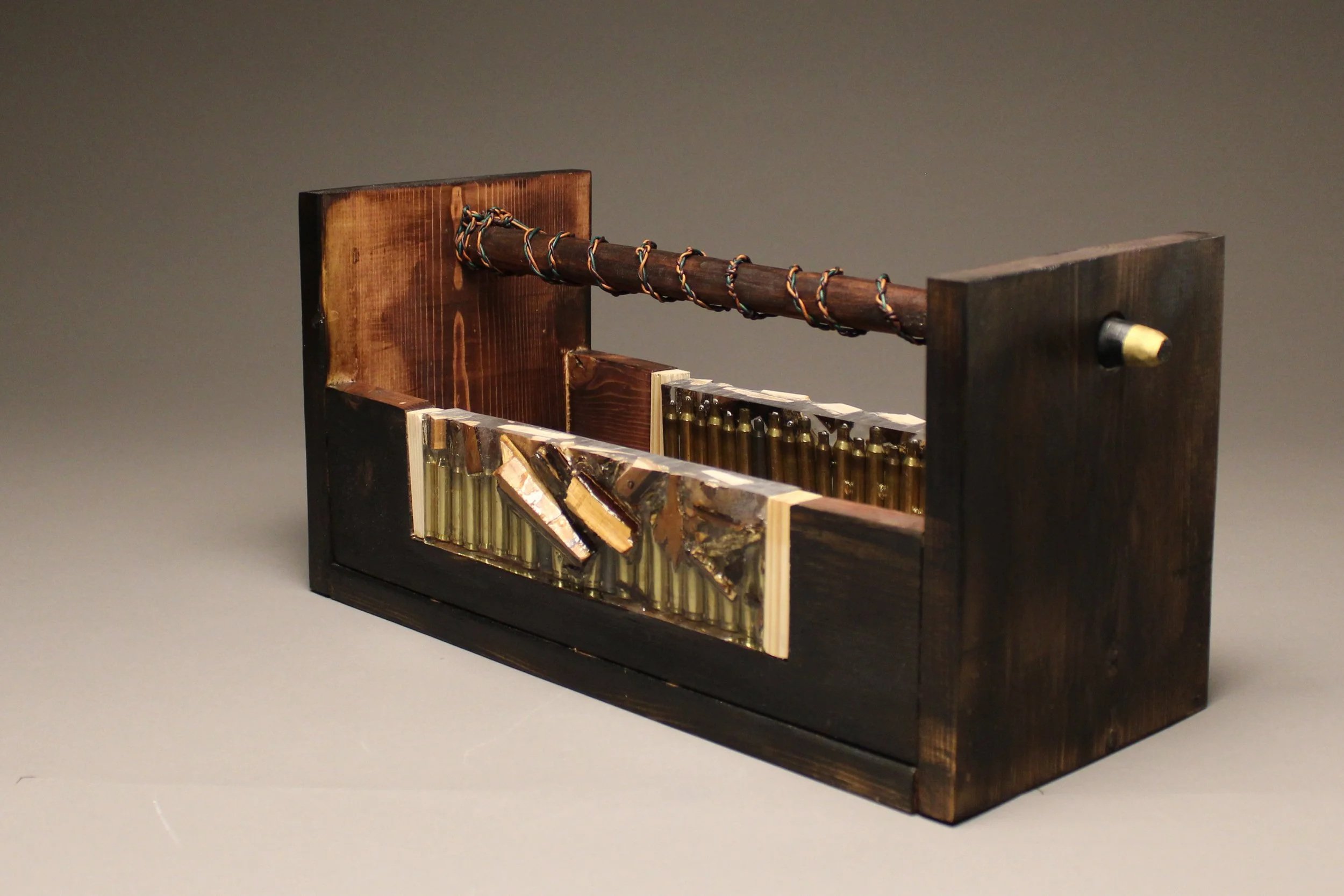A mixed-media art sculpture resembling a rifle, made from various wood pieces, metal, and other materials, with a wooden handle and multiple small metallic parts, set against a plain background.