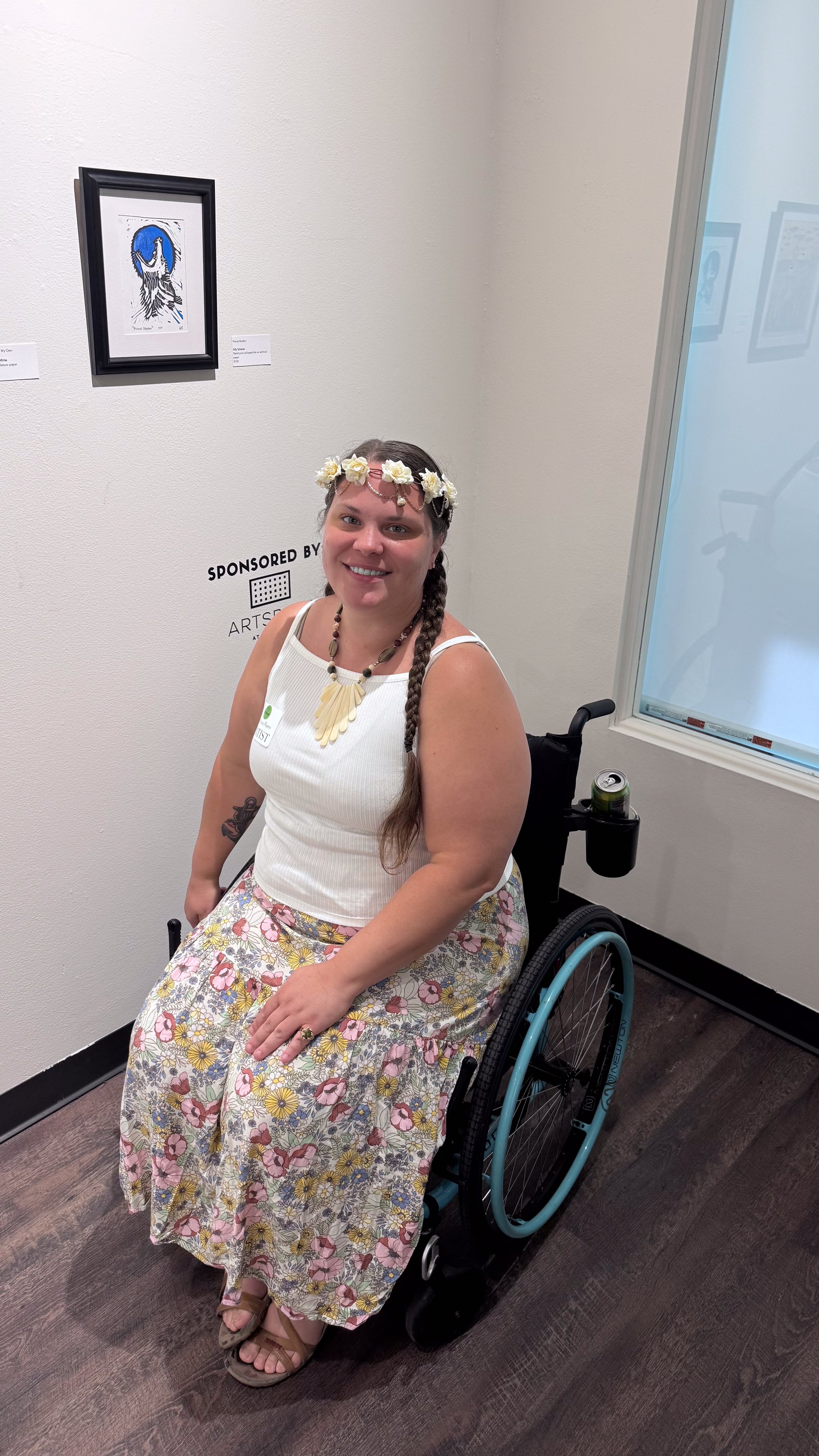 Smiling woman in a wheelchair wearing a floral skirt, white tank top, floral crown, and jewelry in an art gallery with framed artwork on the wall.