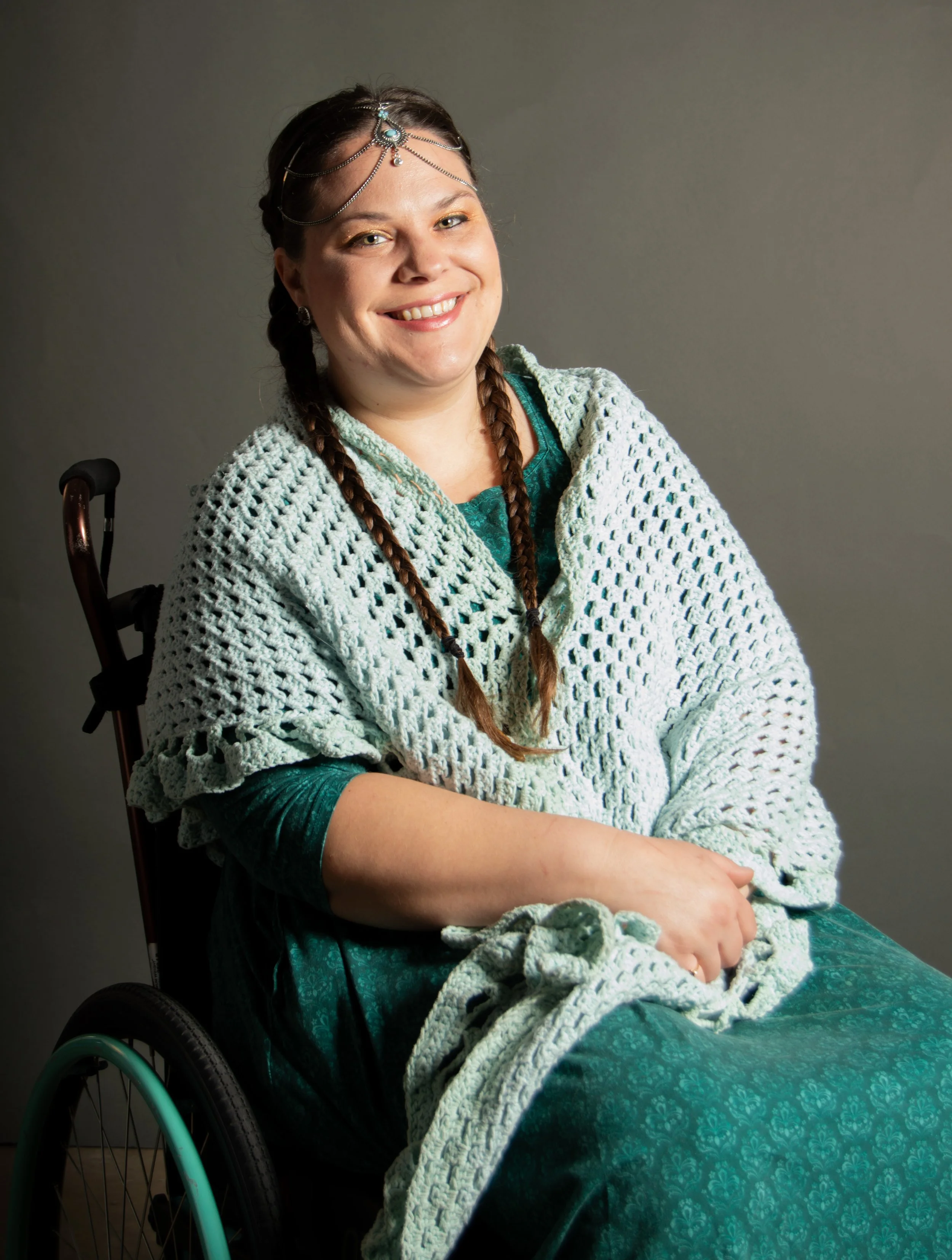 A woman in a wheelchair smiling, wearing a turquoise dress and a white crocheted shawl, with braided hair and jewelry on her forehead.