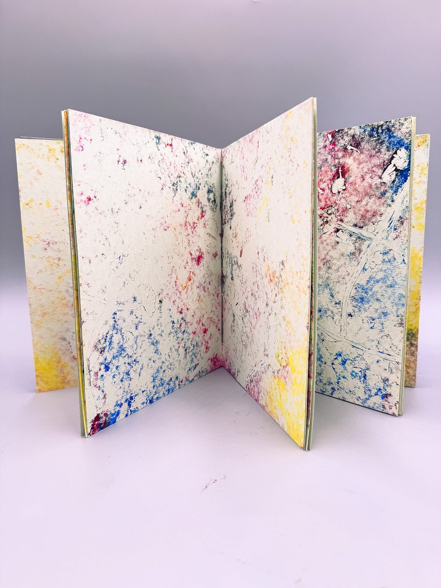 An open, small art book with abstract colorful splatter designs on its pages, standing upright on a white surface against a light purple background.