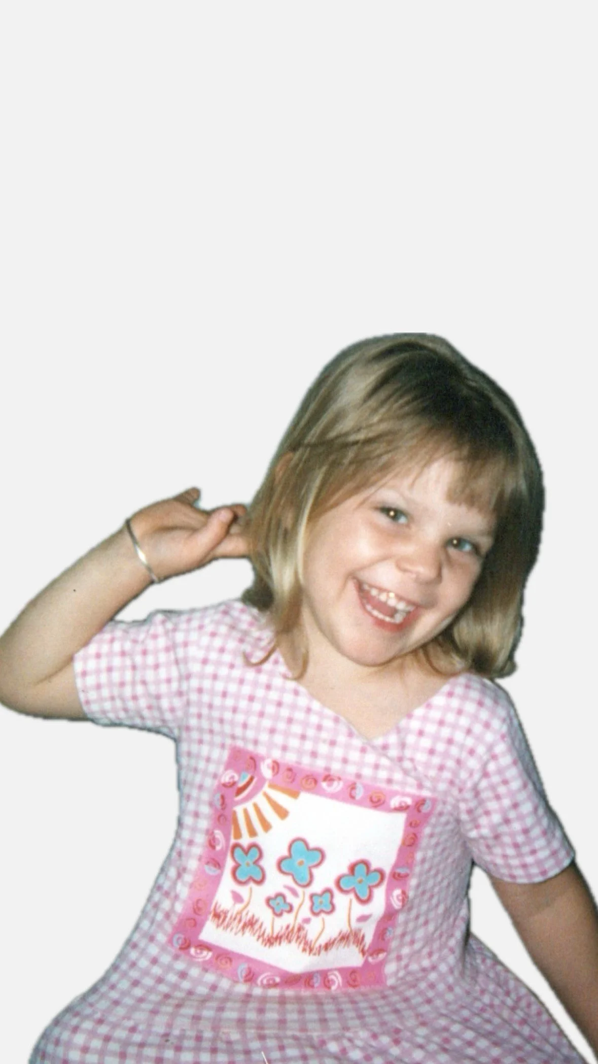 A young girl with light brown hair and a wide smile, wearing a pink and white checkered dress, posing with her right hand behind her head, against a plain background.