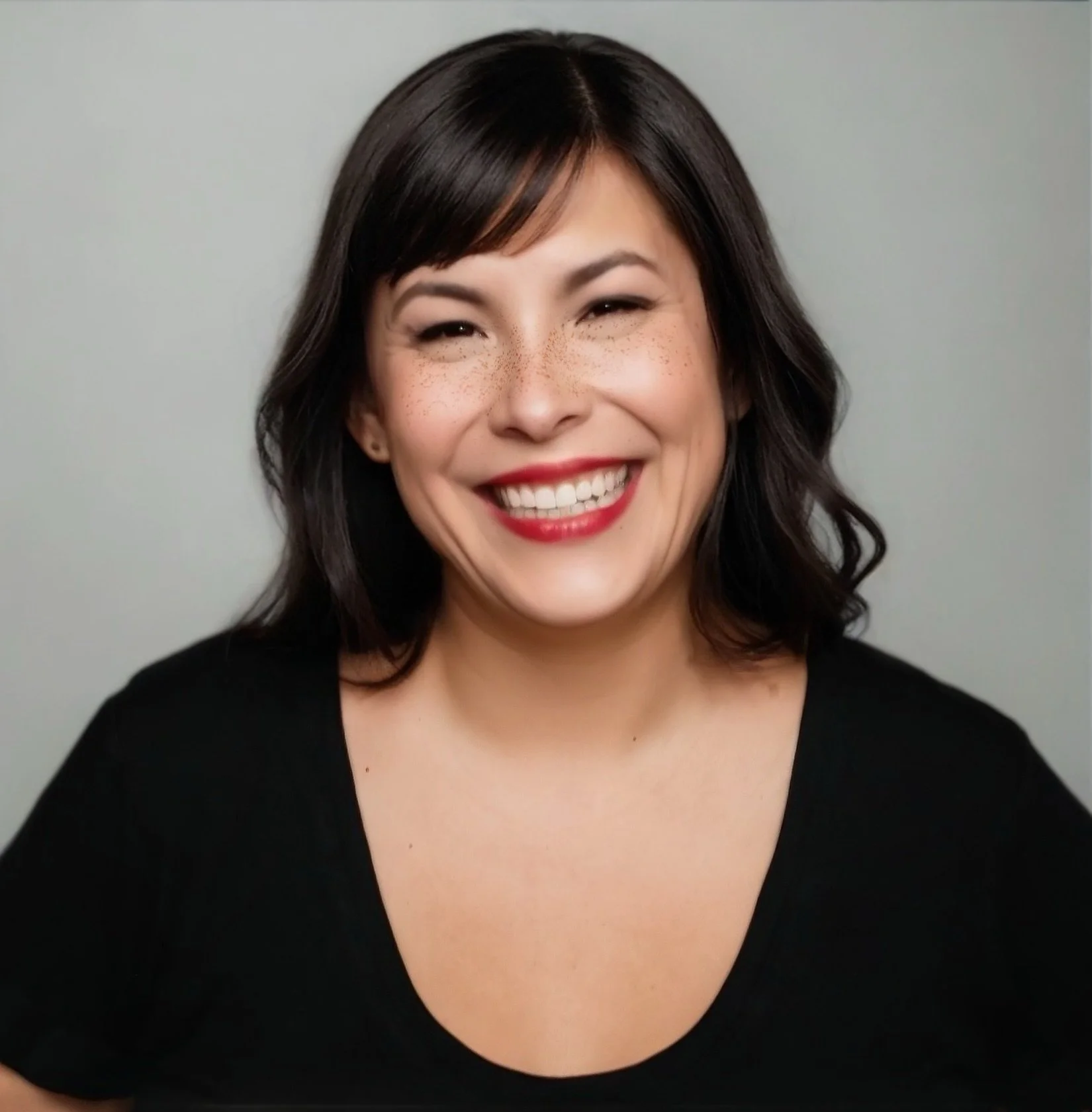 A woman with shoulder-length dark hair smiling with red lipstick and wearing a black top on a gray background.