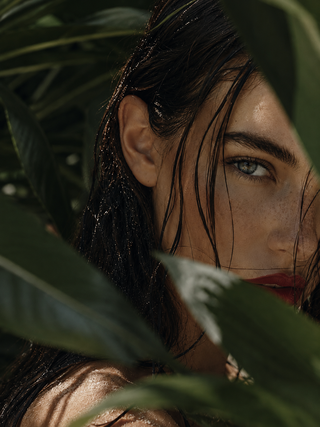 Close-up of a woman's face partially hidden by green leaves, with dark wet hair and piercing blue eyes.