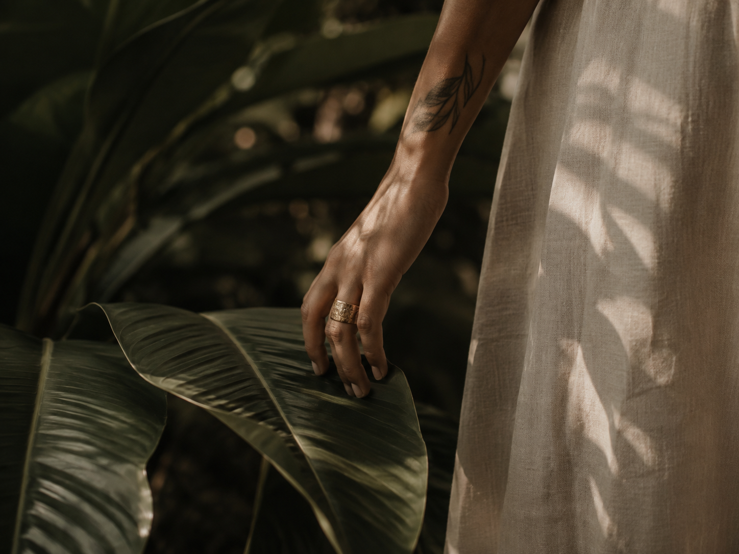 Close-up of a person's hand with a tattoo, wearing a gold ring, gently touching a large green leaf. The person is wearing a light-colored, semi-sheer fabric dress, and there is foliage in the background.
