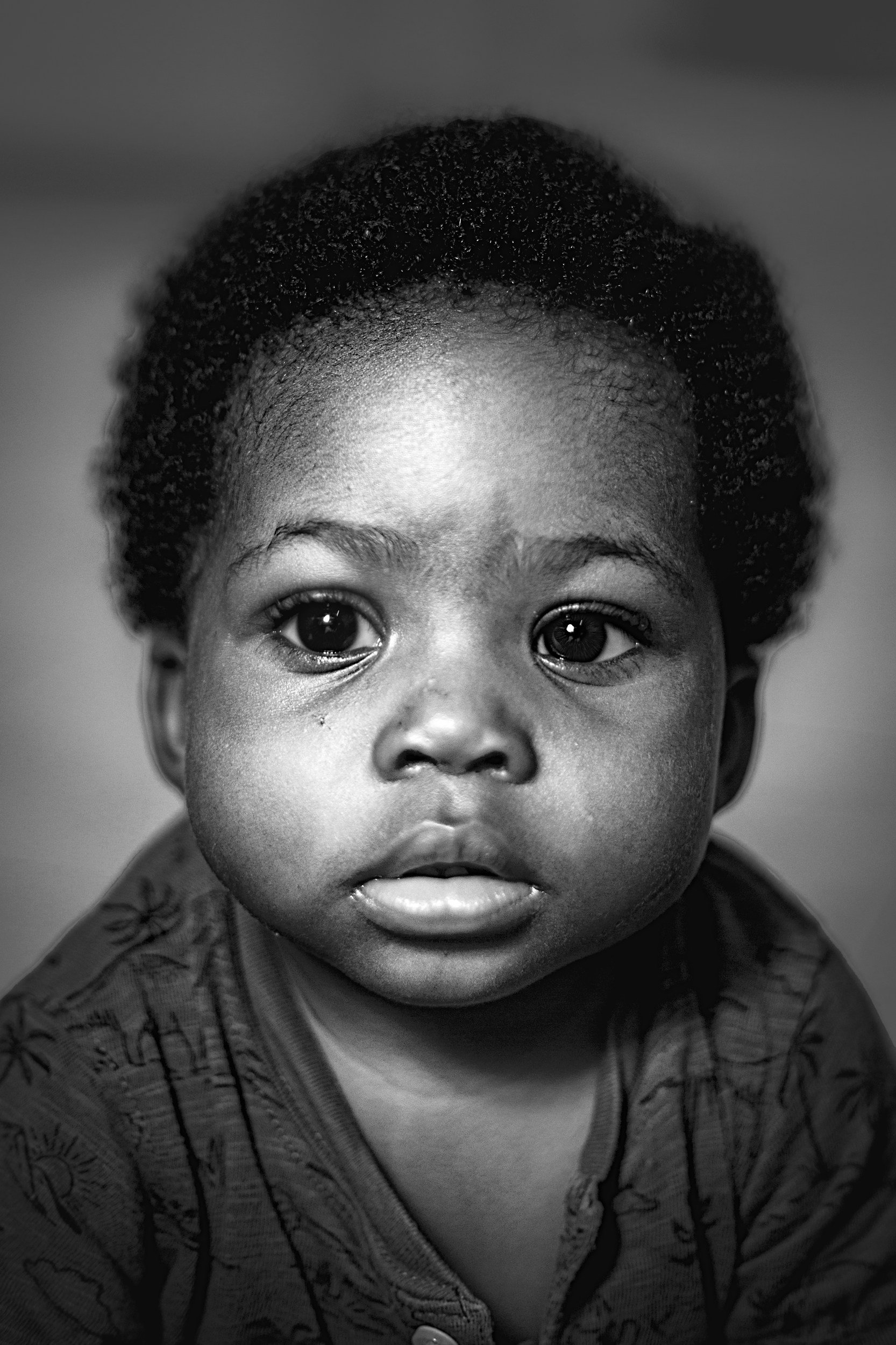 Close-up black and white portrait of a young child with short, curly hair and large, expressive eyes, wearing a patterned hoodie.