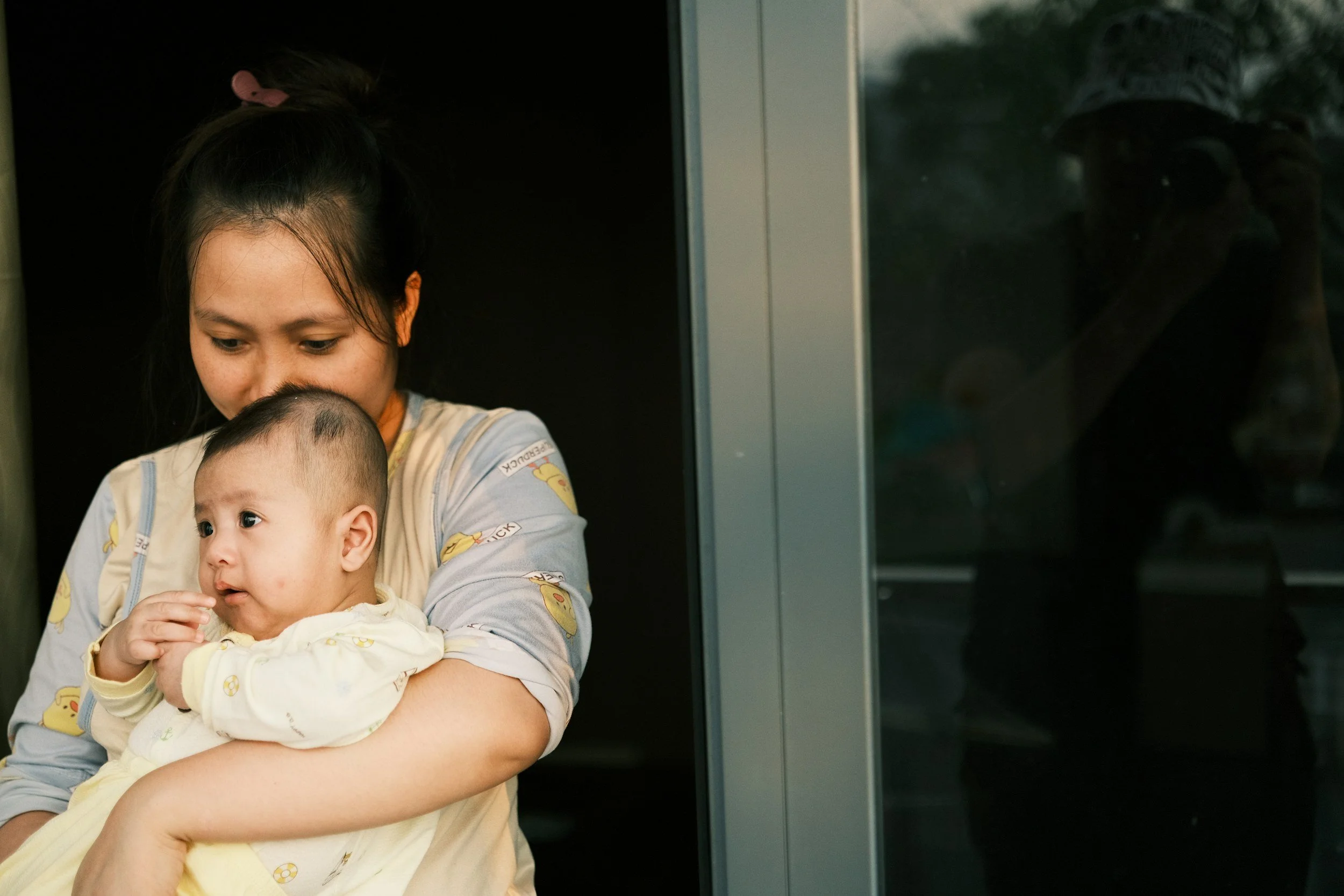 A woman holding a young child while standing near a glass door, with a dog visible outside.