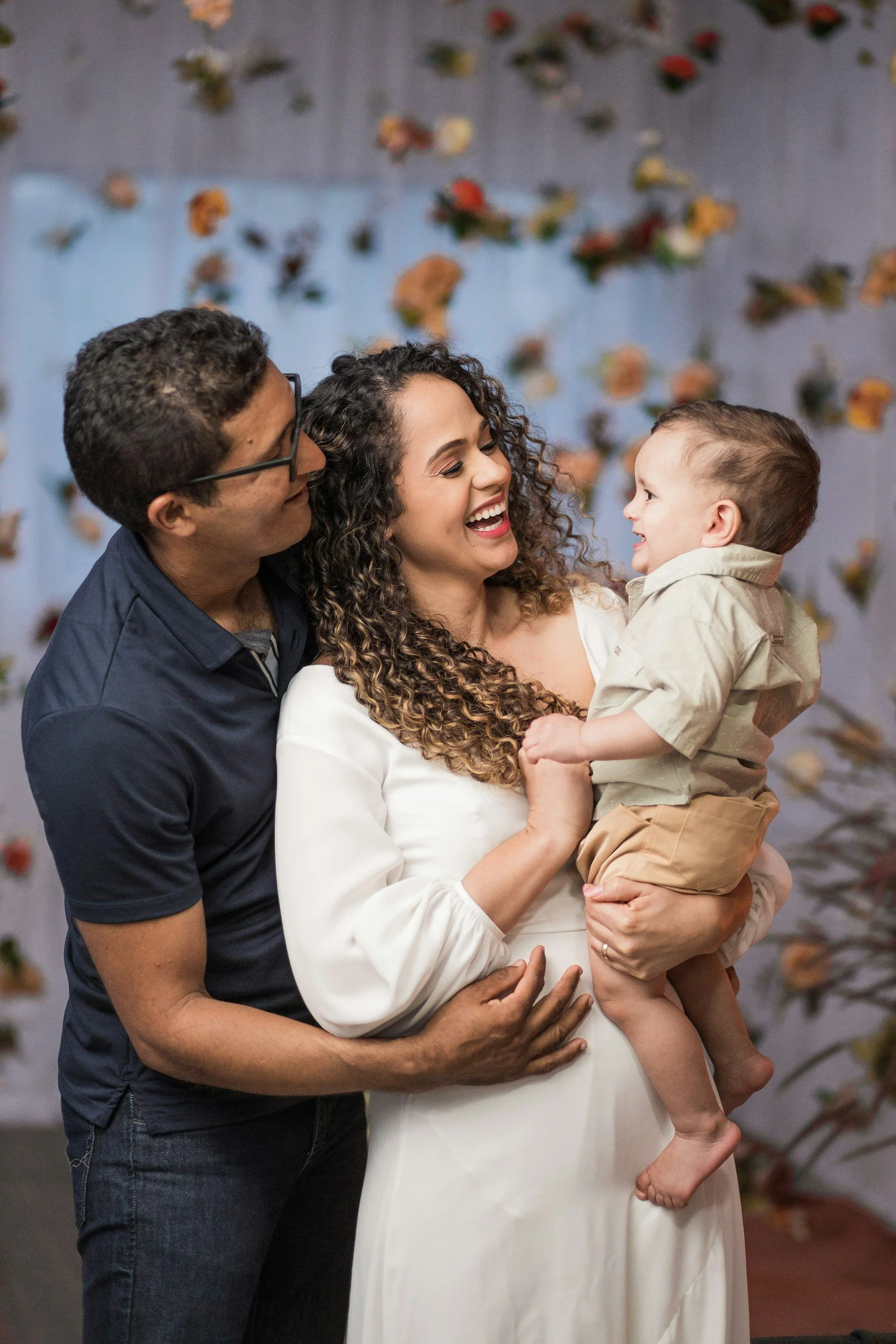 A woman in a white dress holding a young boy with curly hair, a man in glasses and a dark shirt appears to be smiling and looking at them. The background has a wall decorated with scattered leaves.